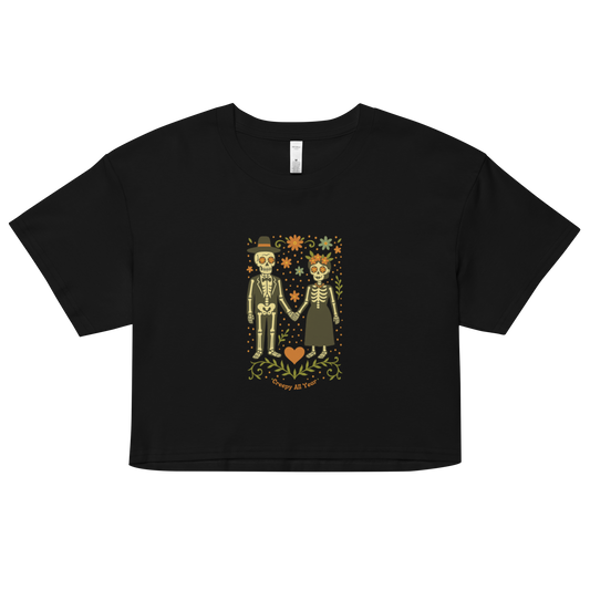 This is a black crop top with a Día de los Muertos–style illustration of a male and female skeleton holding hands, surrounded by marigolds, vines, and folk art patterns. The skeletons wear traditional black clothing, the man in a suit and hat, the woman with a floral crown with a red heart and leaves at the bottom and the text Creepy All Year. Rendered in warm tones of orange, green, and cream, symbolizing eternal love and remembrance 