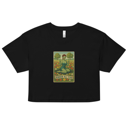 This is a black crop top with a design illustration of a sassy zombie pinup girl standing in a floral field, wearing tattered lingerie and flipping off the viewer with both hands. She has green skin, vintage curls, and a bold attitude. The design blends horror and humor, with a pastel color palette and creepy cute vibes