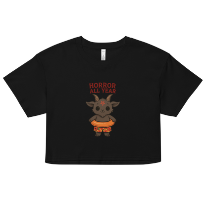 This is a black crop top with a cartoon illustration of a baby Baphomet goat with curved horns and a playful grin, wearing bright floral swim trunks while floating in a colorful inner tube. The character is set against a light background with the bold text “Horror All Year” above in red letters