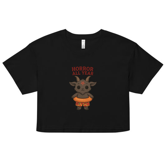 This is a black crop top with a cartoon illustration of a baby Baphomet goat with curved horns and a playful grin, wearing bright floral swim trunks while floating in a colorful inner tube. The character is set against a light background with the bold text “Horror All Year” above in red letters