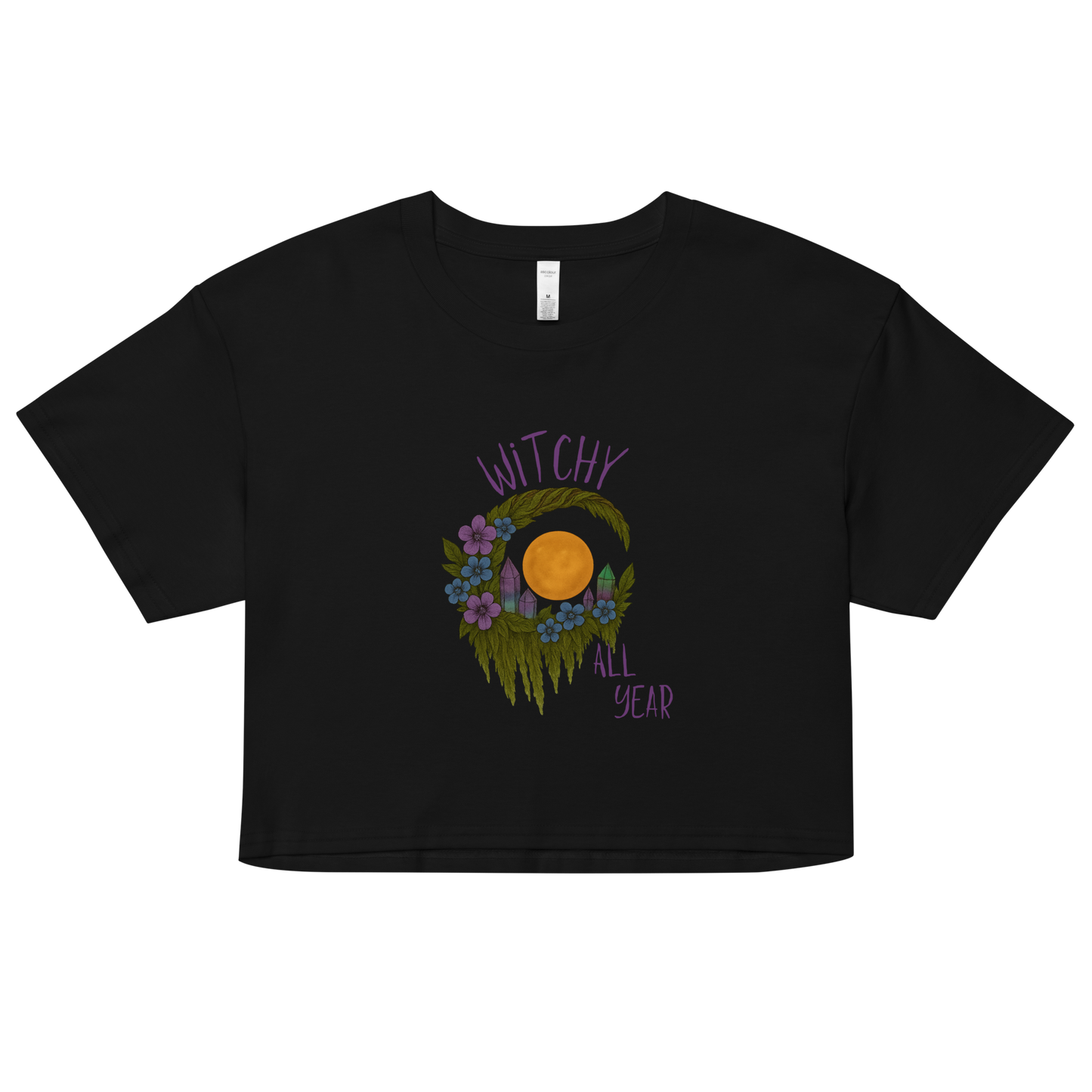 This is a black crop top with a design of a hand-drawn wreath made of moss-covered branches wraps around a glowing sun. The wreath is decorated with purple and blue flowers, small amethyst and fluorite crystals, and strands of hanging moss. The words “Witchy” curve above the wreath and “All Year” sits on the bottom right 
