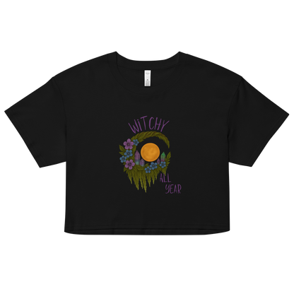This is a black crop top with a design of a hand-drawn wreath made of moss-covered branches wraps around a glowing sun. The wreath is decorated with purple and blue flowers, small amethyst and fluorite crystals, and strands of hanging moss. The words “Witchy” curve above the wreath and “All Year” sits on the bottom right 

