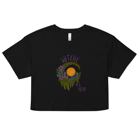 This is a black crop top with a design of a hand-drawn wreath made of moss-covered branches wraps around a glowing sun. The wreath is decorated with purple and blue flowers, small amethyst and fluorite crystals, and strands of hanging moss. The words “Witchy” curve above the wreath and “All Year” sits on the bottom right 
