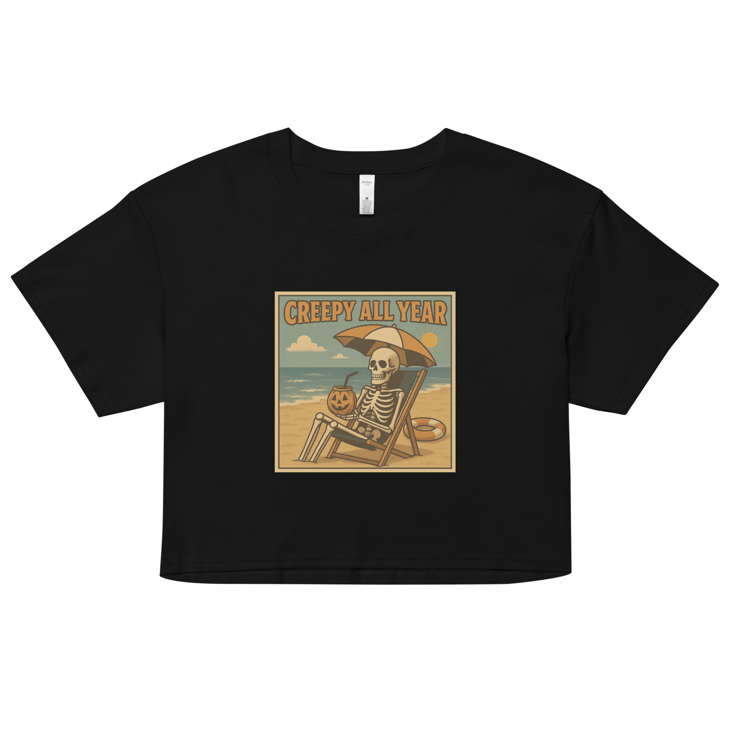 This is a black crop top with a retro-style illustration of a grinning skeleton lounging on a beach chair under an orange umbrella. The skeleton is holding a pumpkin-shaped drink cup with a straw and relaxing by the ocean, with a bright sun in the sky and a life preserver in the sand. The words “Creepy All Year” appear boldly at the top