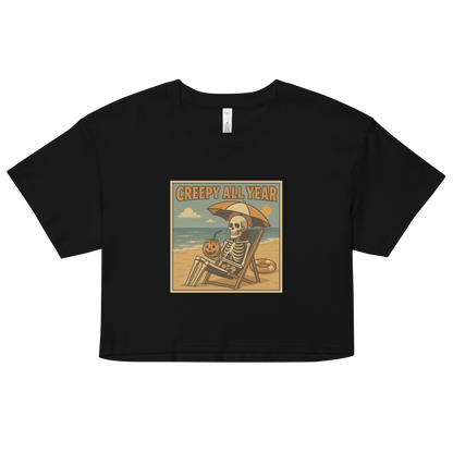 This is a black crop top with a retro-style illustration of a grinning skeleton lounging on a beach chair under an orange umbrella. The skeleton is holding a pumpkin-shaped drink cup with a straw and relaxing by the ocean, with a bright sun in the sky and a life preserver in the sand. The words “Creepy All Year” appear boldly at the top