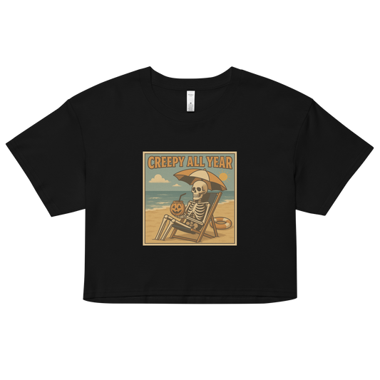 This is a black crop top with a retro-style illustration of a grinning skeleton lounging on a beach chair under an orange umbrella. The skeleton is holding a pumpkin-shaped drink cup with a straw and relaxing by the ocean, with a bright sun in the sky and a life preserver in the sand. The words “Creepy All Year” appear boldly at the top