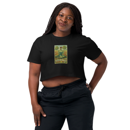 This is a woman with dark braids wearing a black crop top with a design illustration of a sassy zombie pinup girl standing in a floral field, wearing tattered lingerie and flipping off the viewer with both hands. She has green skin, vintage curls, and a bold attitude. The design blends horror and humor, with a pastel color palette and creepy cute vibes