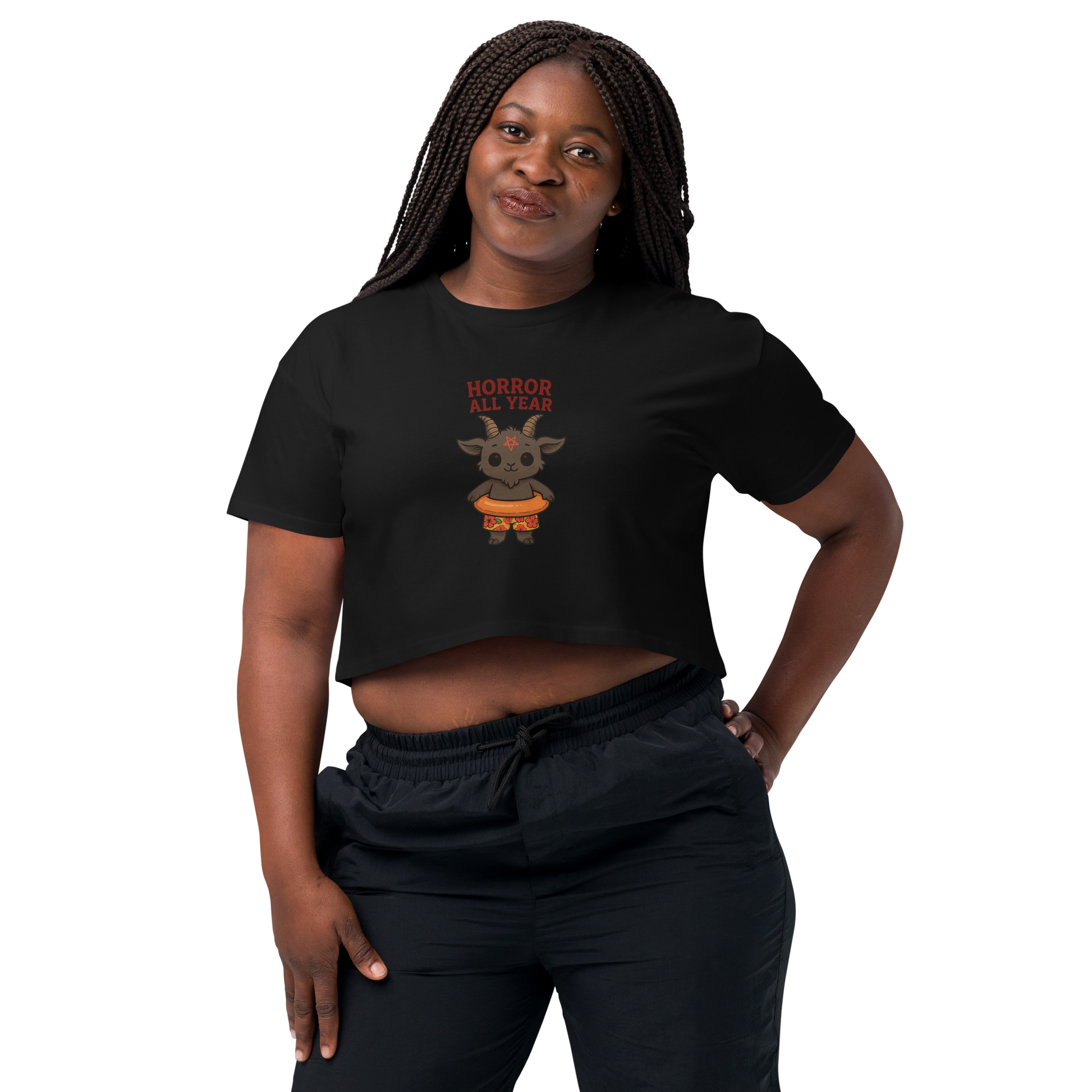 This is a woman with dark braids wearing a black crop top with a cartoon illustration of a baby Baphomet goat with curved horns and a playful grin, wearing bright floral swim trunks while floating in a colorful inner tube. The character is set against a light background with the bold text “Horror All Year” above in red letters