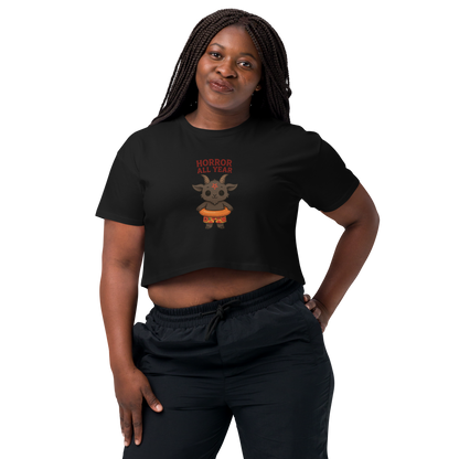 This is a woman with dark braids wearing a black crop top with a cartoon illustration of a baby Baphomet goat with curved horns and a playful grin, wearing bright floral swim trunks while floating in a colorful inner tube. The character is set against a light background with the bold text “Horror All Year” above in red letters