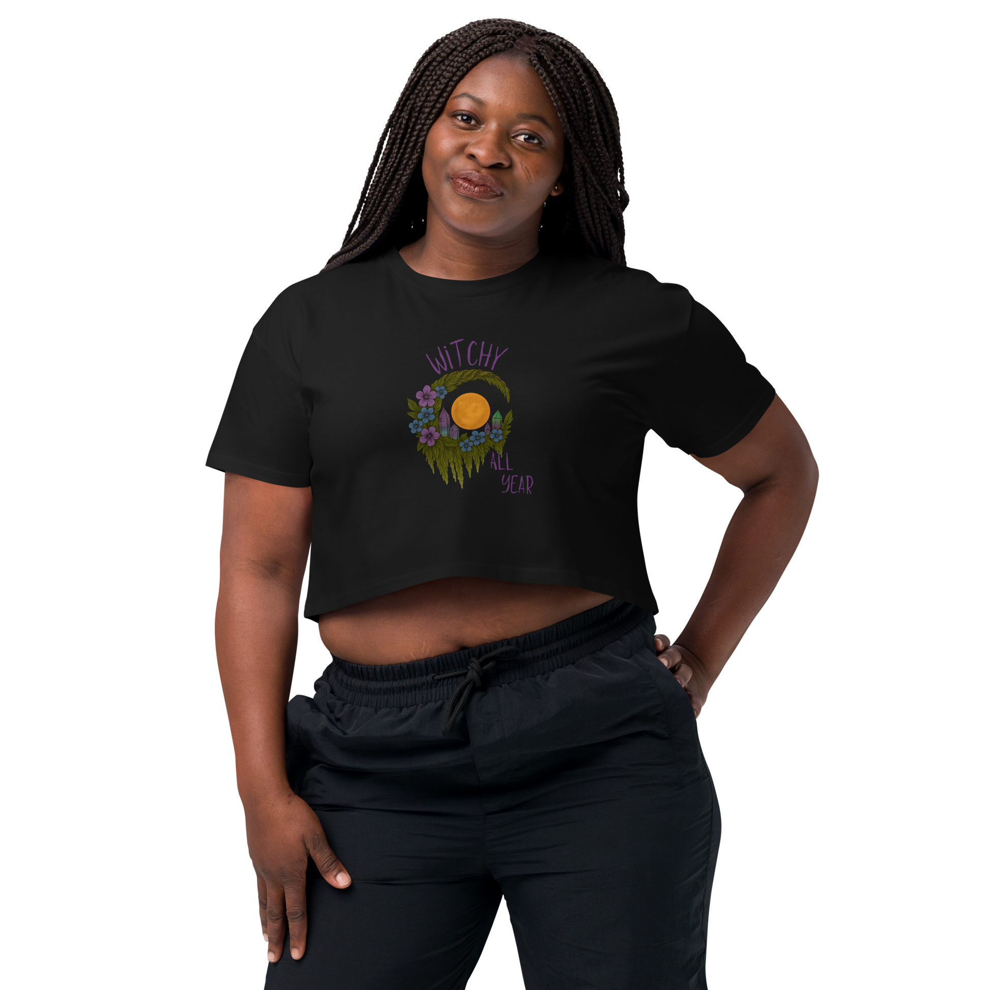 This is a woman with dark braids wearing a black crop top with a design of a hand-drawn wreath made of moss-covered branches wraps around a glowing sun. The wreath is decorated with purple and blue flowers, small amethyst and fluorite crystals, and strands of hanging moss. The words “Witchy” curve above the wreath and “All Year” sits on the bottom right 
