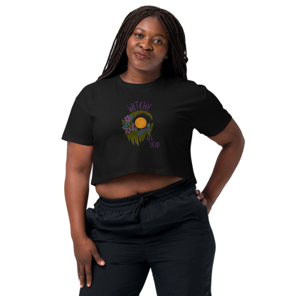 This is a woman with dark braids wearing a black crop top with a design of a hand-drawn wreath made of moss-covered branches wraps around a glowing sun. The wreath is decorated with purple and blue flowers, small amethyst and fluorite crystals, and strands of hanging moss. The words “Witchy” curve above the wreath and “All Year” sits on the bottom right 
