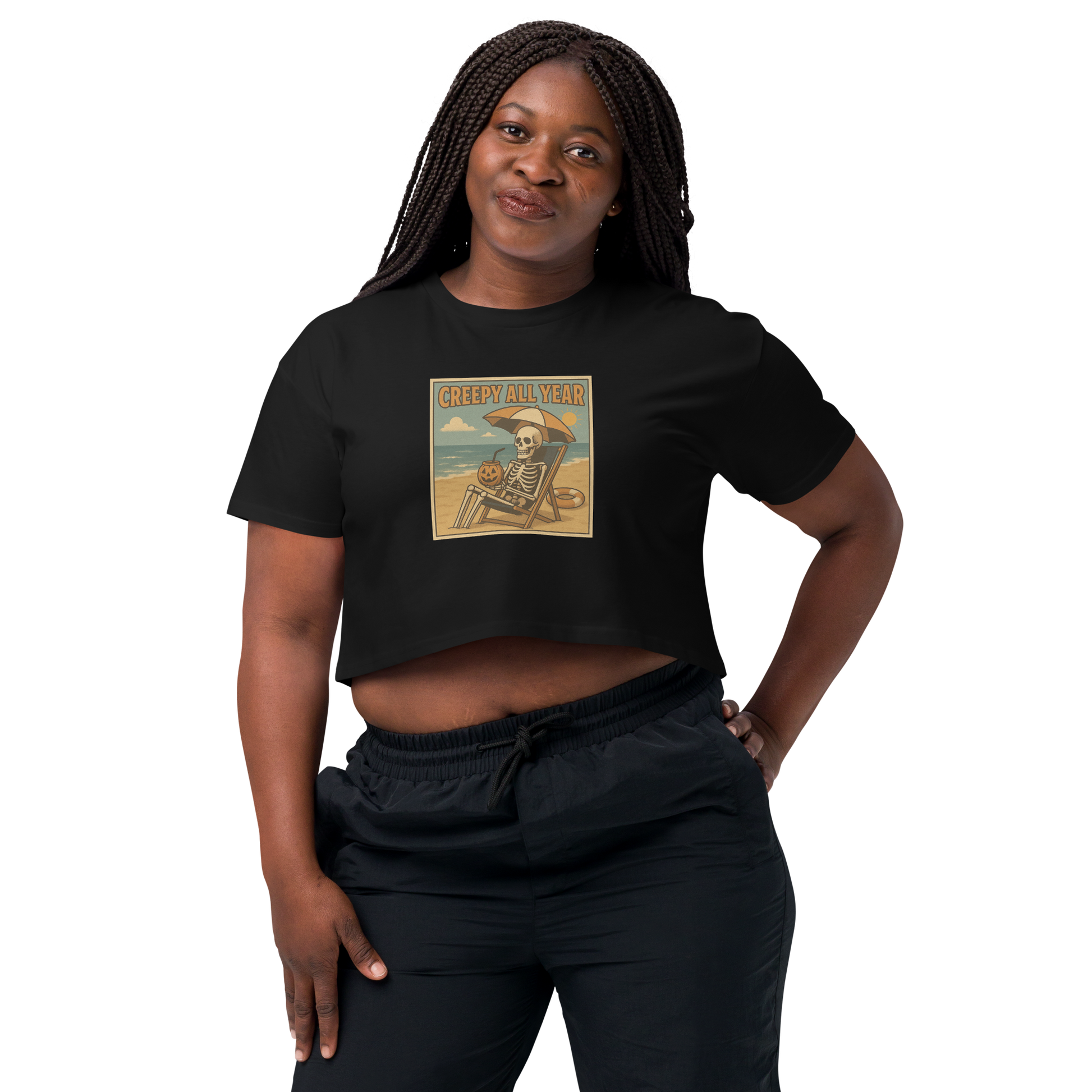 This is a woman with black braids wearing black crop top with a retro-style illustration of a grinning skeleton lounging on a beach chair under an orange umbrella. The skeleton is holding a pumpkin-shaped drink cup with a straw and relaxing by the ocean, with a bright sun in the sky and a life preserver in the sand. The words “Creepy All Year” appear boldly at the top 