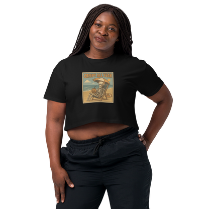 This is a woman with black braids wearing black crop top with a retro-style illustration of a grinning skeleton lounging on a beach chair under an orange umbrella. The skeleton is holding a pumpkin-shaped drink cup with a straw and relaxing by the ocean, with a bright sun in the sky and a life preserver in the sand. The words “Creepy All Year” appear boldly at the top 