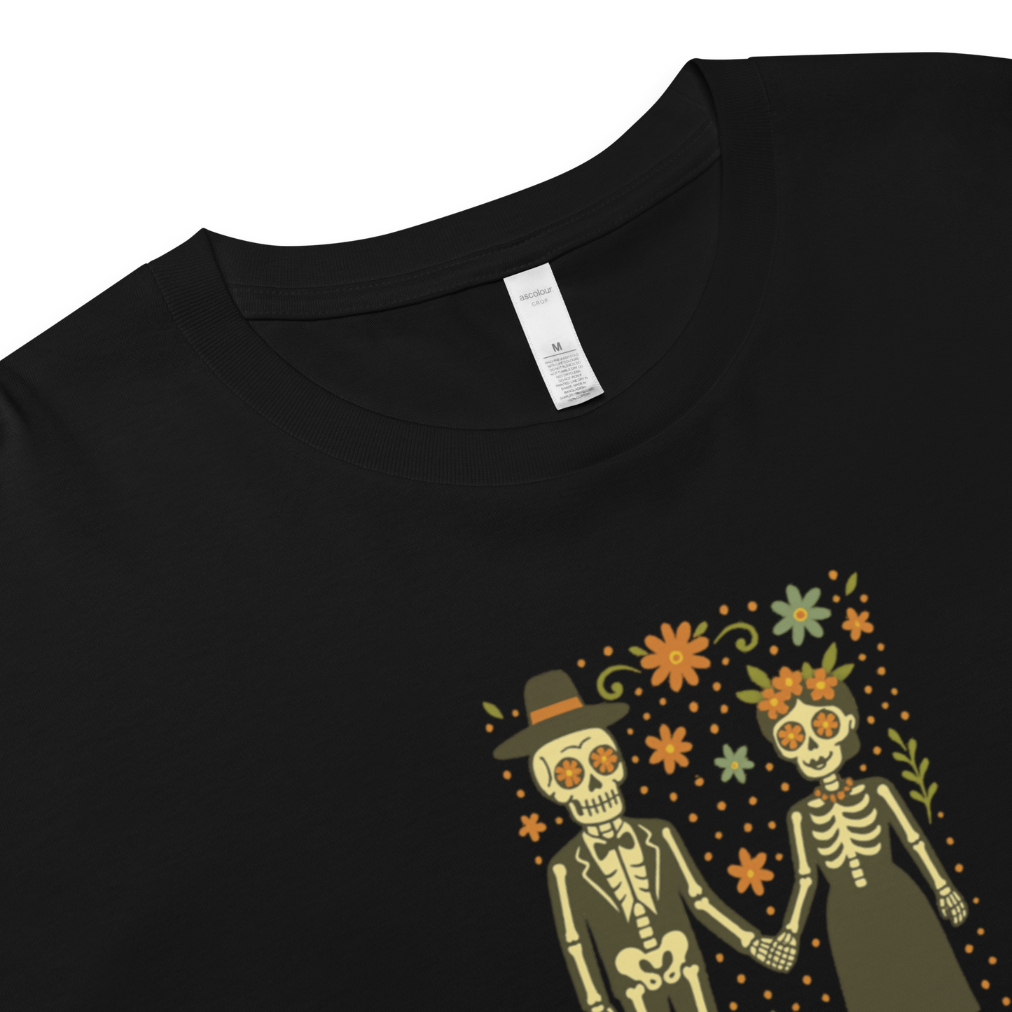 This is a zoom in on a black crop top with a Día de los Muertos–style illustration of a male and female skeleton holding hands, surrounded by marigolds, vines, and folk art patterns. The skeletons wear traditional black clothing, the man in a suit and hat, the woman with a floral crown with a red heart and leaves at the bottom and the text Creepy All Year. Rendered in warm tones of orange, green, and cream, symbolizing eternal love and remembrance