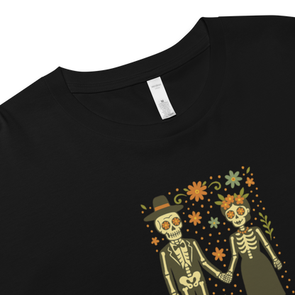 This is a zoom in on a black crop top with a Día de los Muertos–style illustration of a male and female skeleton holding hands, surrounded by marigolds, vines, and folk art patterns. The skeletons wear traditional black clothing, the man in a suit and hat, the woman with a floral crown with a red heart and leaves at the bottom and the text Creepy All Year. Rendered in warm tones of orange, green, and cream, symbolizing eternal love and remembrance
