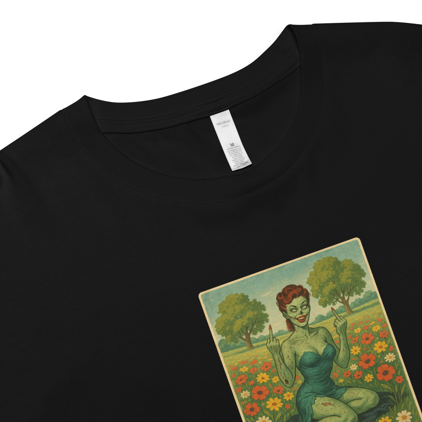 This is a zoom in on a black crop top with a design illustration of a sassy zombie pinup girl standing in a floral field, wearing tattered lingerie and flipping off the viewer with both hands. She has green skin, vintage curls, and a bold attitude. The design blends horror and humor, with a pastel color palette and creepy cute vibes