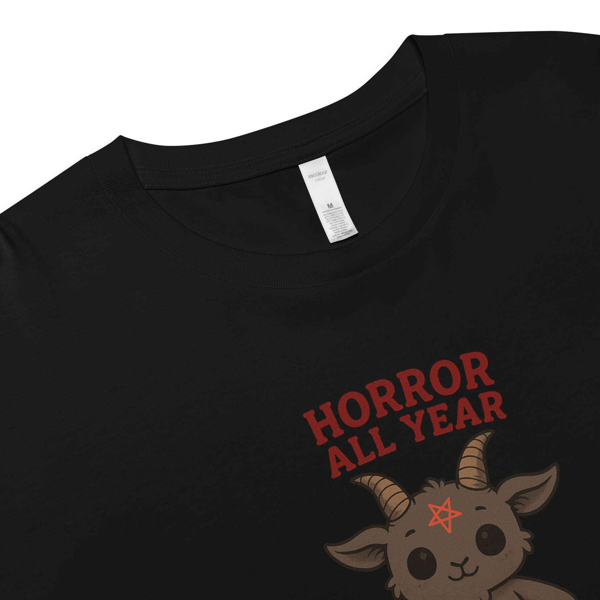 This is a zoom in of a black crop top with a cartoon illustration of a baby Baphomet goat with curved horns and a playful grin, wearing bright floral swim trunks while floating in a colorful inner tube. The character is set against a light background with the bold text “Horror All Year” above in red letters