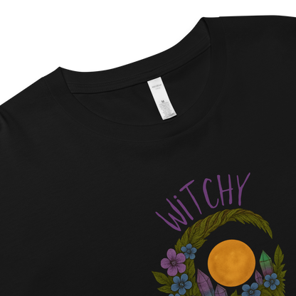 This is a zoom in of a black crop top with a design of a hand-drawn wreath made of moss-covered branches wraps around a glowing sun. The wreath is decorated with purple and blue flowers, small amethyst and fluorite crystals, and strands of hanging moss. The words “Witchy” curve above the wreath and “All Year” sits on the bottom right 
