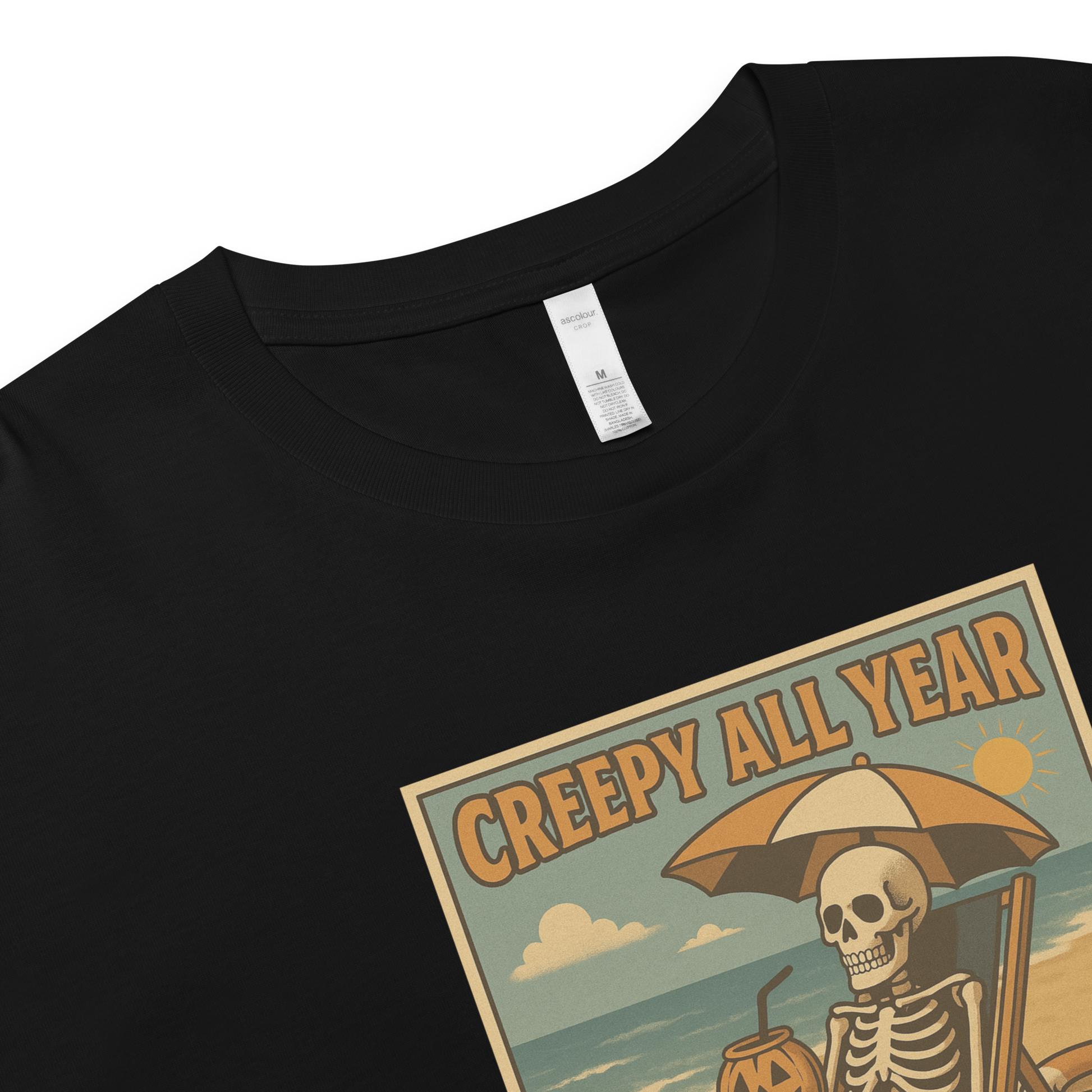 This is a zoomed in black crop top with a retro-style illustration of a grinning skeleton lounging on a beach chair under an orange umbrella. The skeleton is holding a pumpkin-shaped drink cup with a straw and relaxing by the ocean, with a bright sun in the sky and a life preserver in the sand. The words “Creepy All Year” appear boldly at the top 